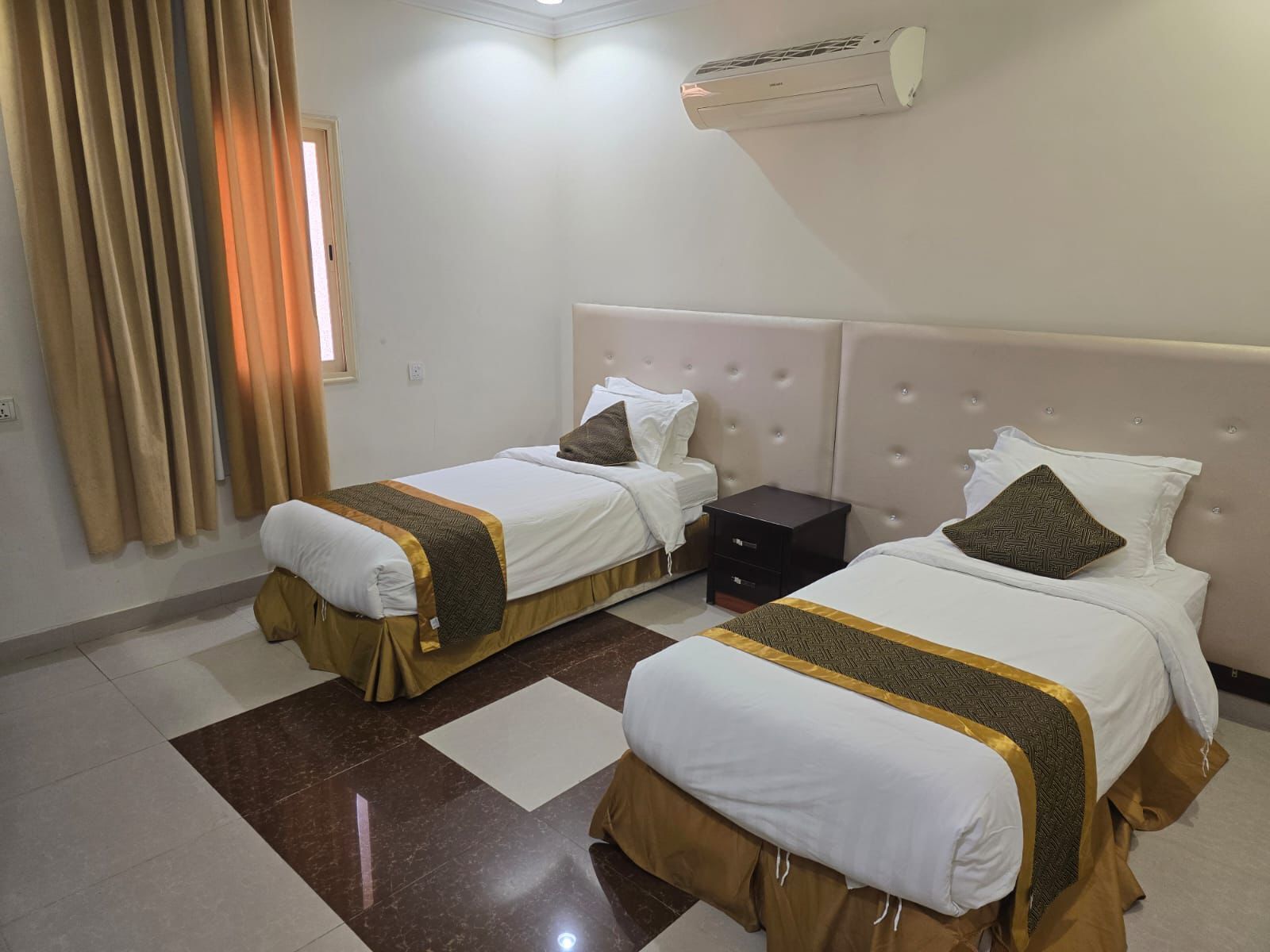 A hotel room with two beds, a wall-mounted air conditioner, and a window with curtains.