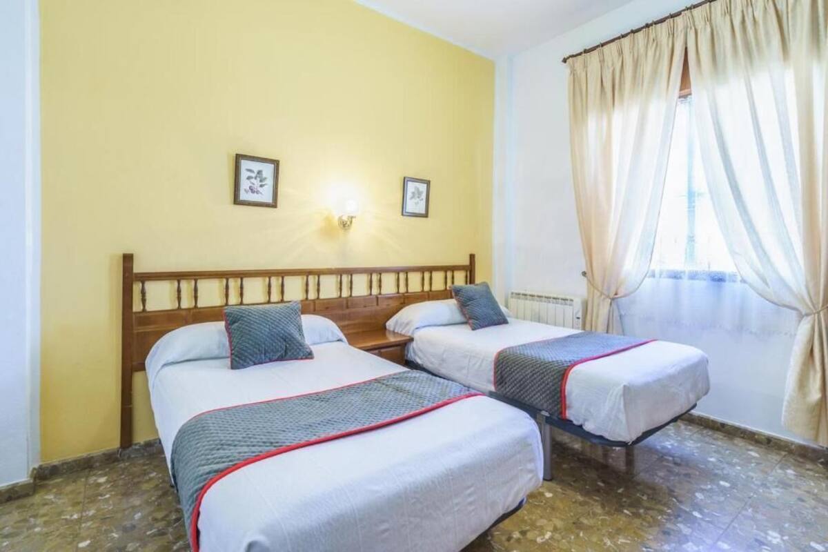 A room with two beds, a wooden headboard, a wall-mounted light, and a window with curtains.