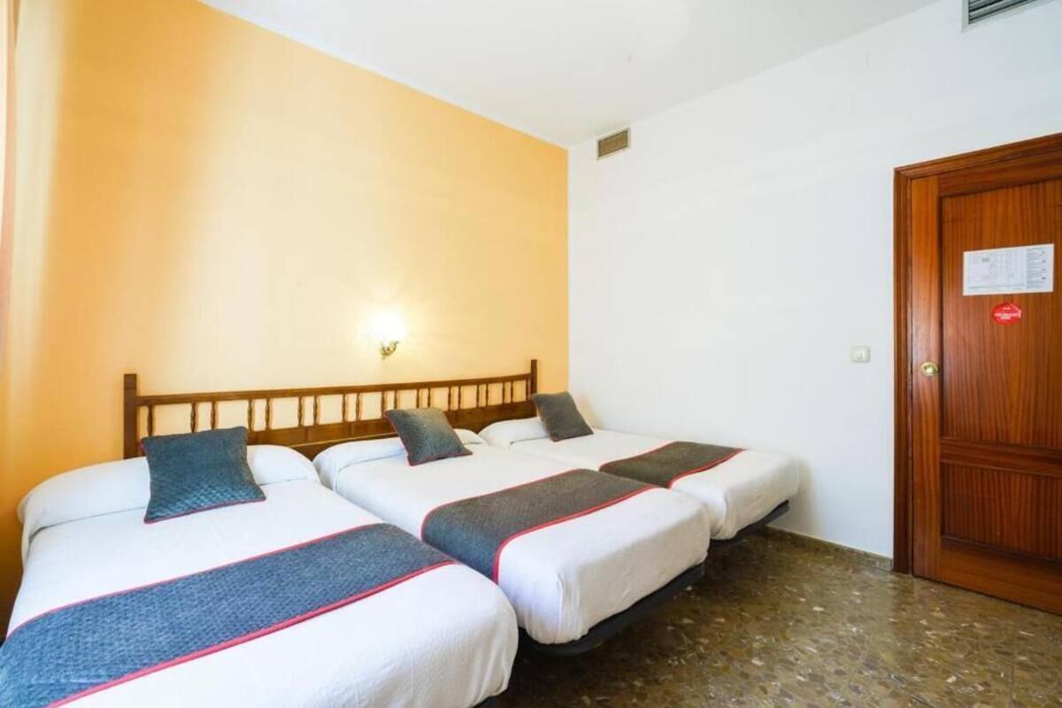 A hotel room with three beds, a wooden headboard, a wall-mounted light, and a wooden door.
