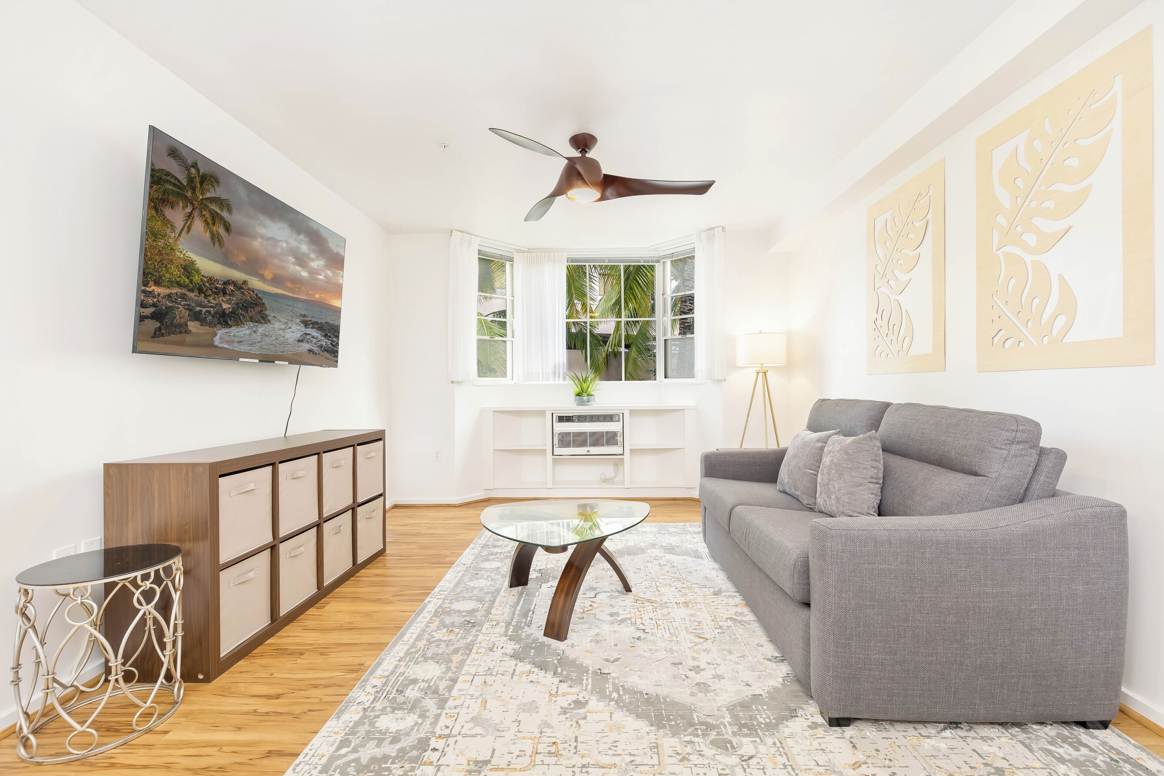 A living room with a grey sofa, a glass coffee table, a wooden cabinet, a ceiling fan, a large TV, and a bay window with a view of the outdoors.