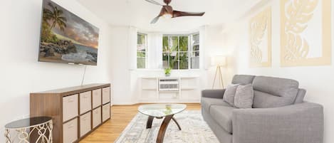 A living room with a grey sofa, a glass coffee table, a wooden cabinet, a ceiling fan, a large TV, and a bay window with a view of the outdoors.