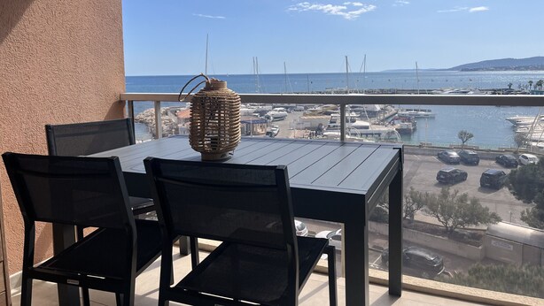Un balcón con mesa y sillas, con vista a un puerto deportivo con barcos y un cielo despejado.