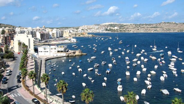 Un pueblo costero con un puerto deportivo lleno de barcos, edificios a lo largo de la costa y un cielo despejado.