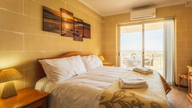 Ein Hotelzimmer mit Bett, Nachttisch mit Lampe, Balkon mit Aussicht und einem Bild an der Wand.