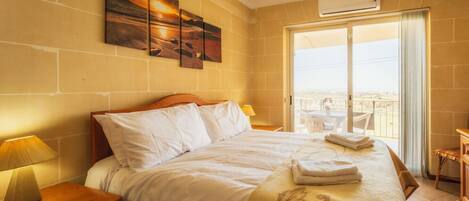 A hotel room with a bed, a nightstand with a lamp, a balcony with a view, and a painting on the wall.