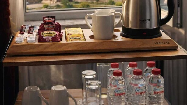 A breakfast setup with a kettle, mug, and snacks on a wooden table, with a view of a castle and mountains through a window. - Ari Otel (Erzurum)