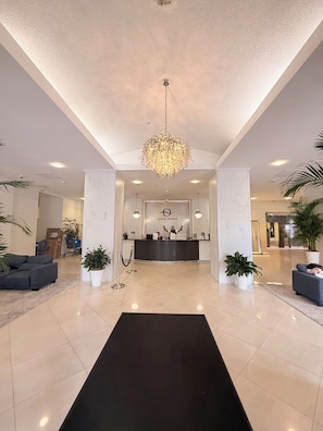 A hotel lobby with a reception desk, a chandelier, potted plants, and a black carpet leading to the desk.