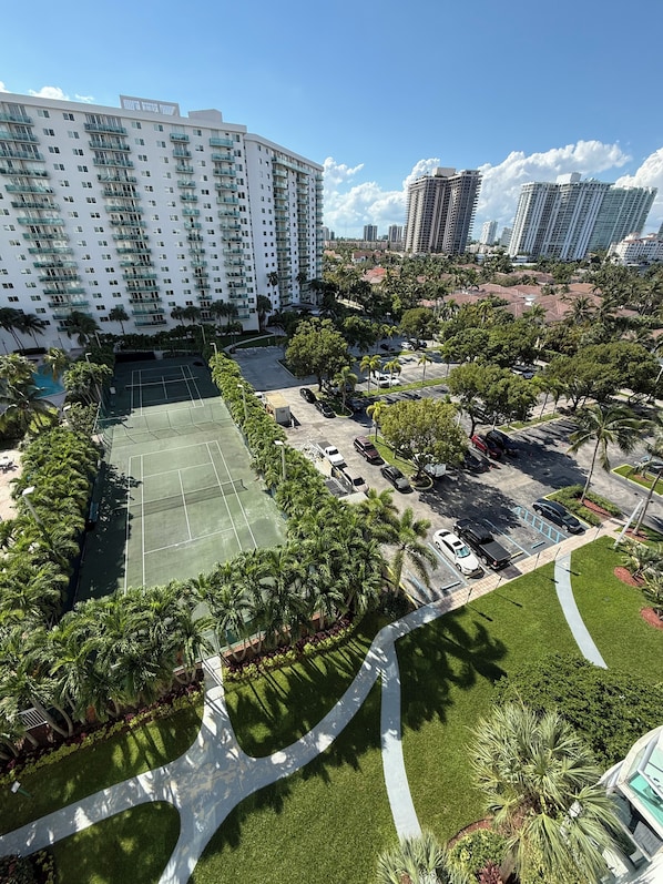 A high-rise hotel with multiple balconies, a tennis court, and a parking area surrounded by palm trees.