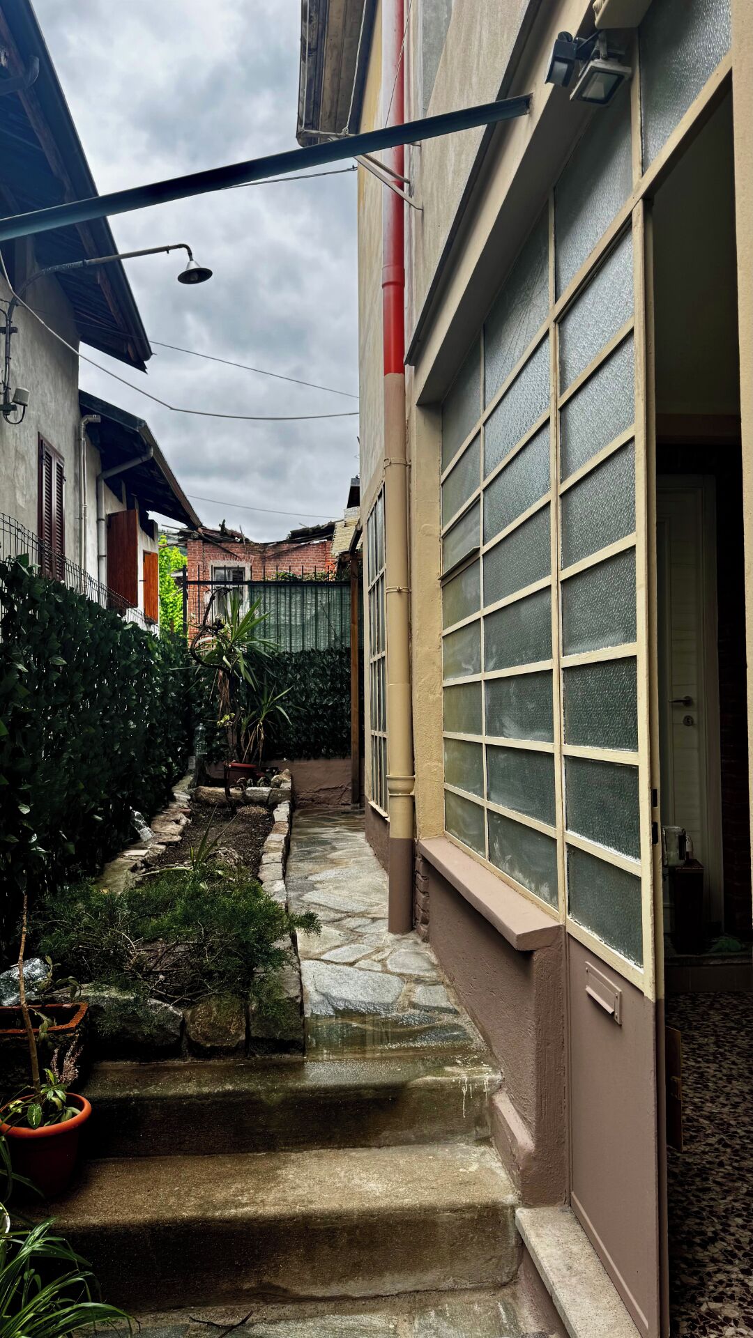 Une ruelle étroite avec des marches menant à l’entrée d’un immeuble, bordée de plantes en pot et d’un chemin pavé.