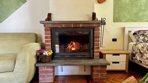 A fireplace with a brick surround, a flower vase on the mantel, a wooden nightstand, a bed with floral bedding, and a sofa. - Charming Apartment with Wood Fireplace, Garden & Mountain View near Oropa (Biella)