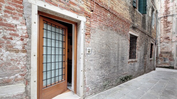 A narrow alleyway with a brick building, a wooden door with a metal grid, and a small window with bars. - ANGELS' HOUSE (Venezia)