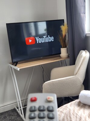 Ein Flachbildfernseher auf einem Holzständer mit einem YouTube-Logo, einer Fernbedienung, einem weißen Stuhl und einem Blumentopf.