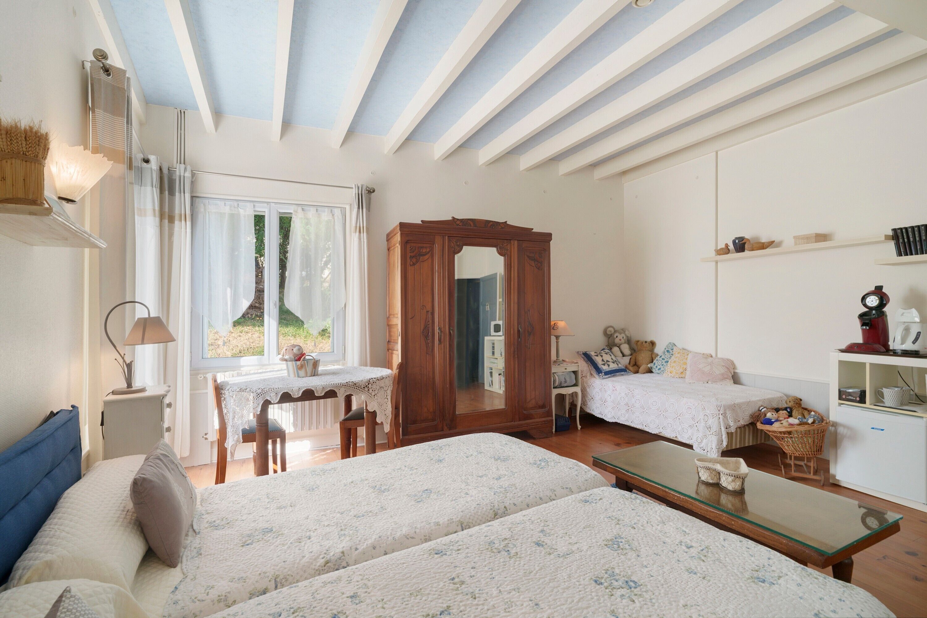 A room with two beds, a wooden wardrobe, a desk with a chair, a small table, and a shelf with books and decorative items.