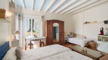A room with two beds, a wooden wardrobe, a desk with a chair, a small table, and a shelf with books and decorative items.