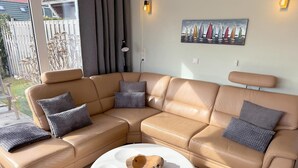 En beige lædersofa i sektioner med grå hynder, et rundt hvidt sofabord med en trædekoration, og et maleri af en sejlbåd på væggen.