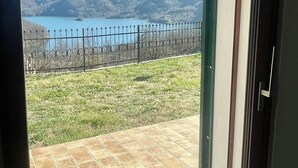 A view through a glass door to a patio with a metal fence, a grassy area, and a lake with mountains in the background. - Casale Nelli - Window on the Lake (Petrella Salto)