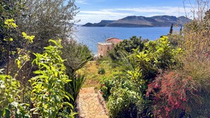 A coastal landscape with a stone pathway leading to a building with a red roof, surrounded by lush greenery and overlooking a body of water. - Villa Maria Peninsula (Kas)