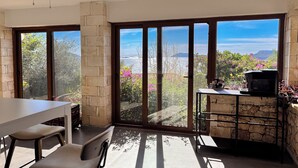 A room with a dining table, chairs, a stone pillar, a fireplace, and large sliding glass doors with a view of the sea and mountains. - Villa Maria Peninsula (Kas)