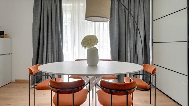 Un comedor con una mesa redonda, sillas naranjas, un jarrón blanco con flores y cortinas grises.