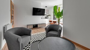 Una sala de estar moderna con un televisor de pantalla plana, dos sillones grises, una mesa de centro redonda y una planta verde grande.