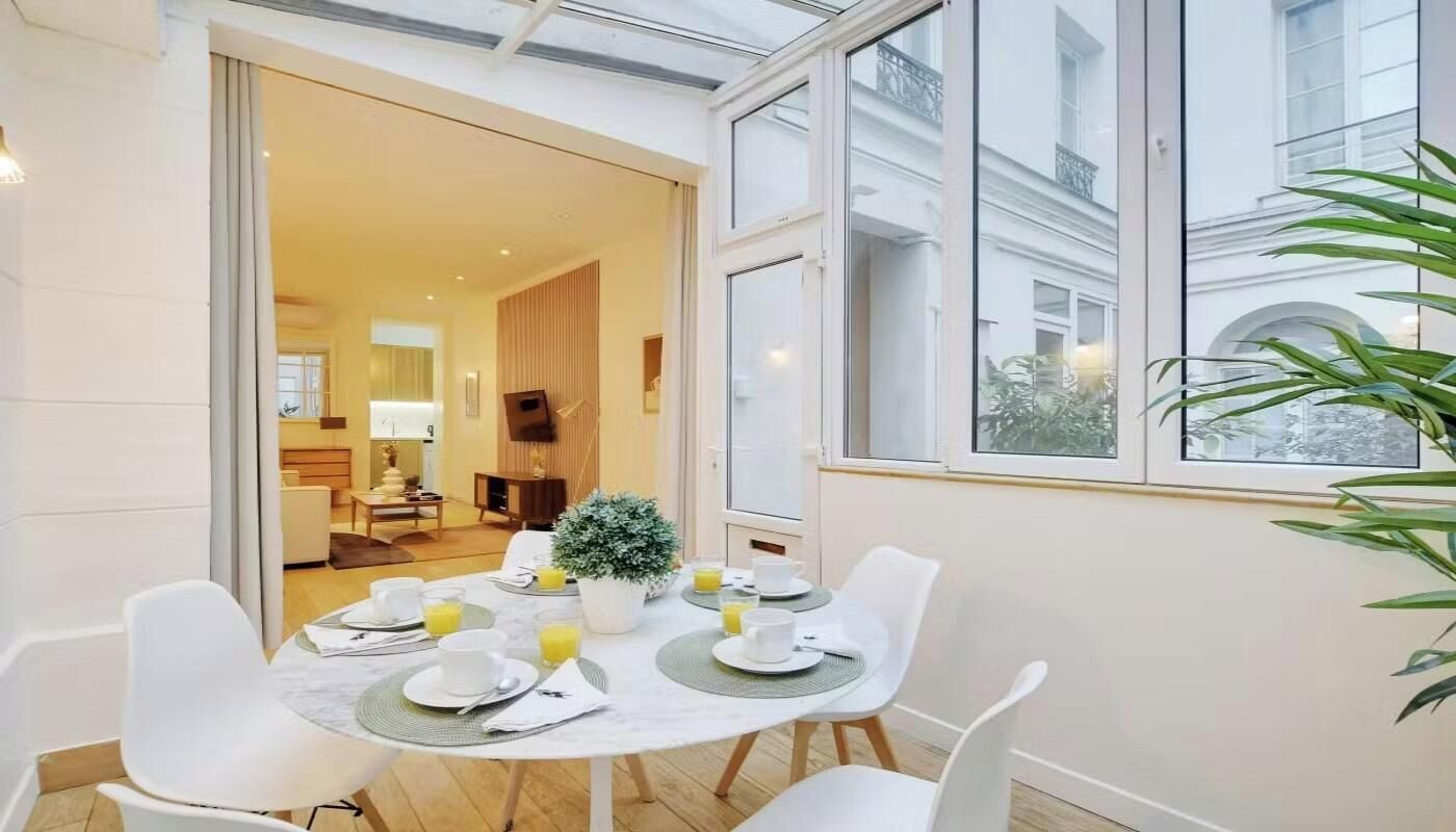 A dining area with a table set for four, a potted plant, and a view of a building through large windows.