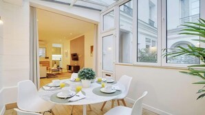 Comedor con una mesa preparada para cuatro, una planta en maceta y vistas a un edificio a través de amplias ventanas.