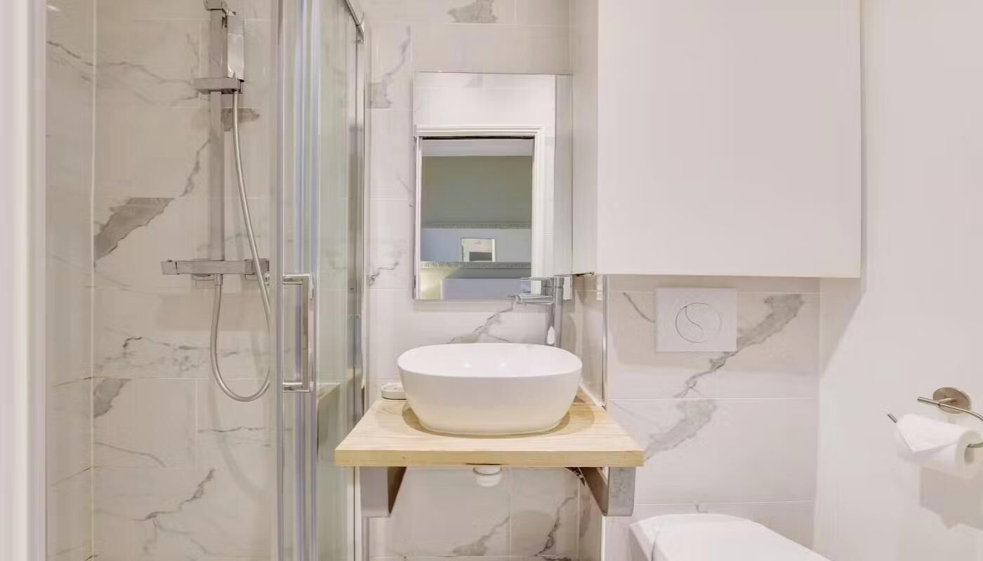 A bathroom with a glass-enclosed shower, a marble-tiled wall, a white sink on a wooden countertop, and a mirror above the sink.