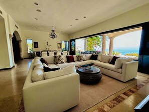 A spacious living room with a large sofa, coffee table, and a view of the ocean. - Luxury 5BD With Stunning Ocean View - Casa Jalisco (San José del Cabo)