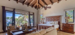 A spacious bedroom with a large bed, a wooden dresser, a seating area with a sofa and chairs, and large windows offering a view of palm trees. - Luxury 5BD With Stunning Ocean View - Casa Jalisco (San José del Cabo)