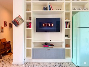 A living room with a TV mounted on a white entertainment unit, a bookshelf, and a teal refrigerator. - Casa Paolo Retro-design With Balcony Near Metro (Milan)