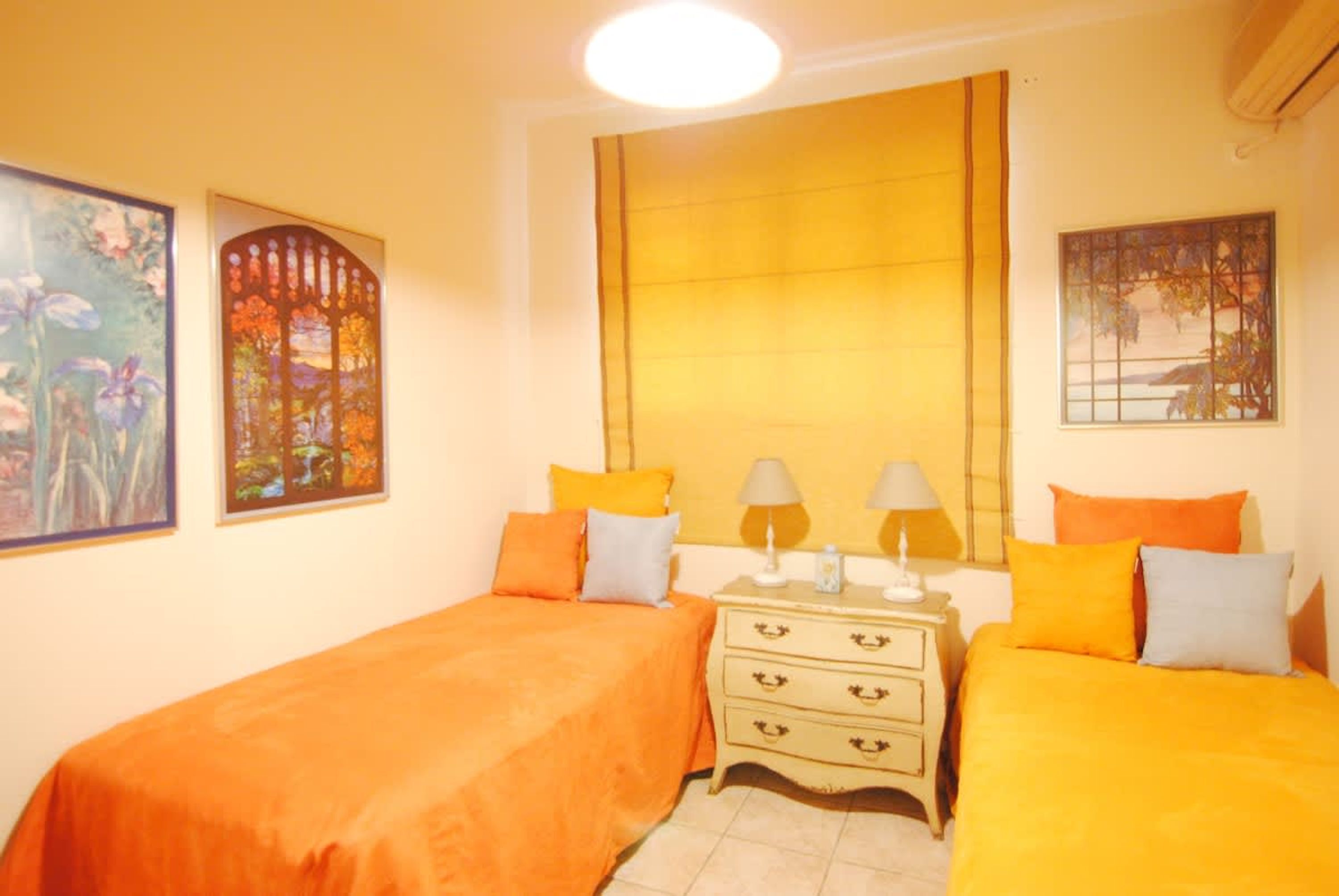 A room with two beds, a nightstand with lamps, and artwork on the walls.