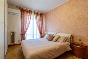 Un dormitorio con cama, cortinas, un radiador, suelo de madera y una mesita de noche.
