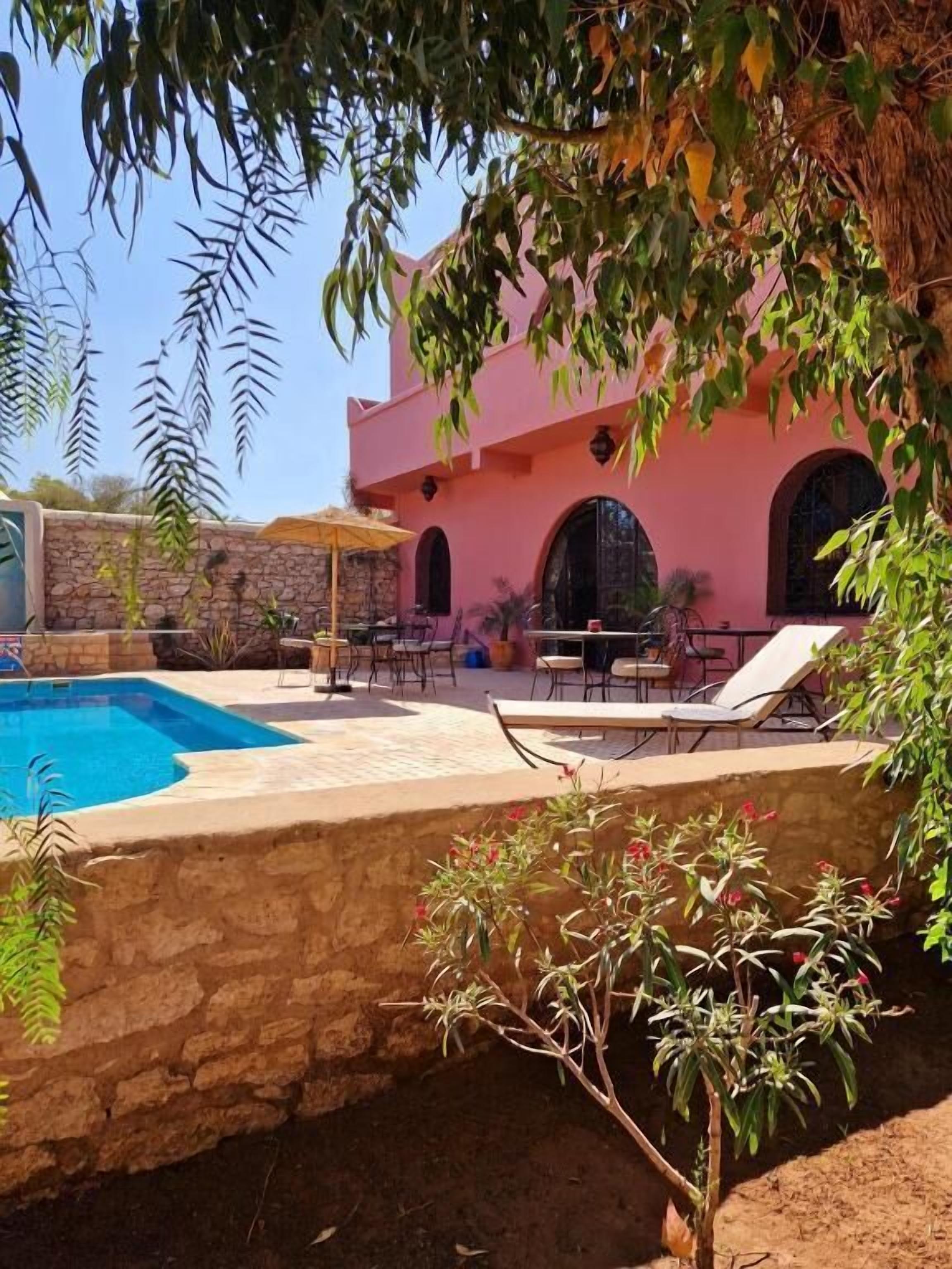 Zona de piscina con sillas de descanso, una pared de piedra y un edificio rosa con ventanas arqueadas.