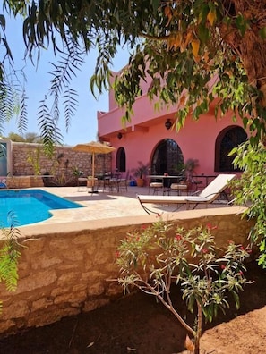 Uma área de piscina com espreguiçadeiras, um muro de pedra e um edifício rosa com janelas em arco.