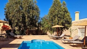 A rectangular swimming pool with a tiled deck, surrounded by lounge chairs and umbrellas, with trees in the background. - Villa Nouran (Ghazoua)