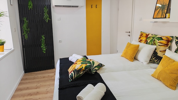 Habitación de hotel con cama, ventana con planta, puerta con una franja vertical y un equipo de aire acondicionado.