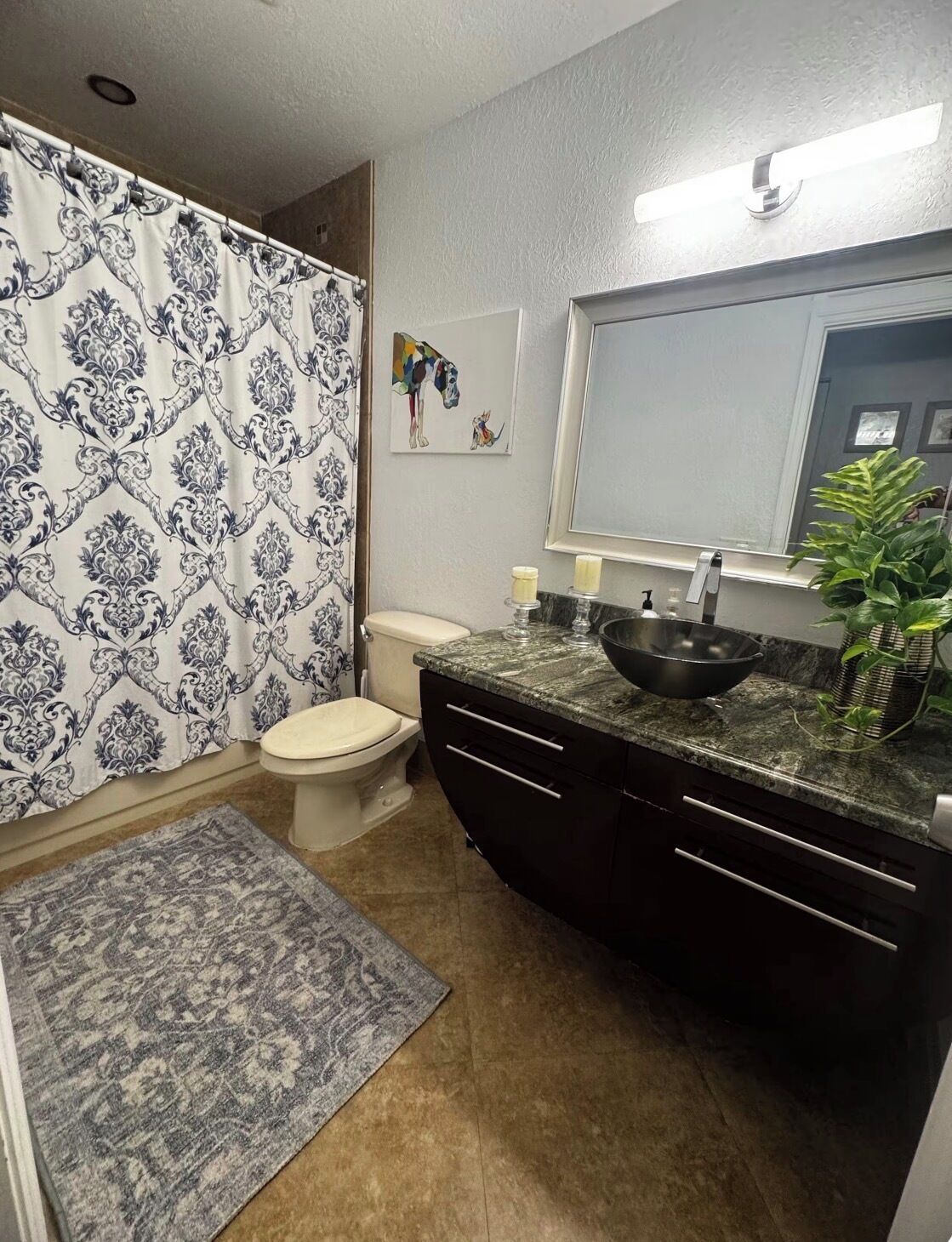 A bathroom with a toilet, a sink with a mirror, a shower curtain, a rug, and a wall-mounted picture.