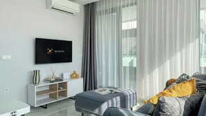 Ein modernes Wohnzimmer mit einem Flachbildfernseher, einer Couch mit gemusterten Kissen, einem Beistelltisch mit Dekoration und transparenten Vorhängen.