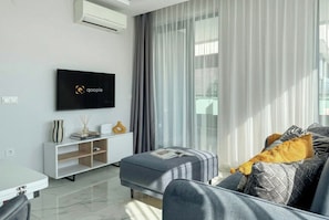 En moderne stue med et fladskærms-tv, en sofa med mønstrede puder, et sofabord med dekorative genstande og lette gardiner.