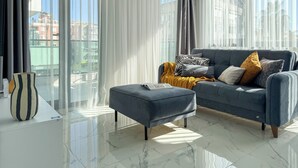 Ein modernes Wohnzimmer mit einem grauen Sofa, passendem Hocker und einem großen Fenster mit transparenten Vorhängen.