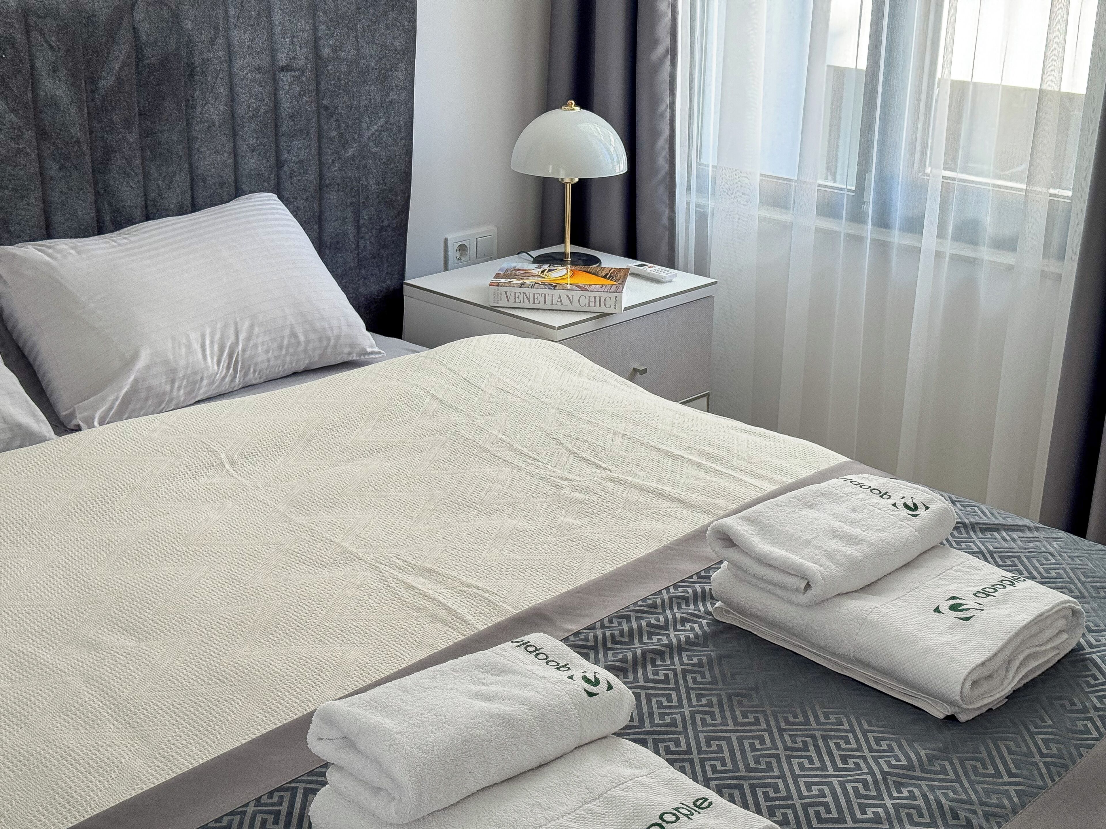 A neatly made bed with a patterned bedspread, a bedside lamp, a book titled 'Venetian Chic,' and a window with sheer curtains.