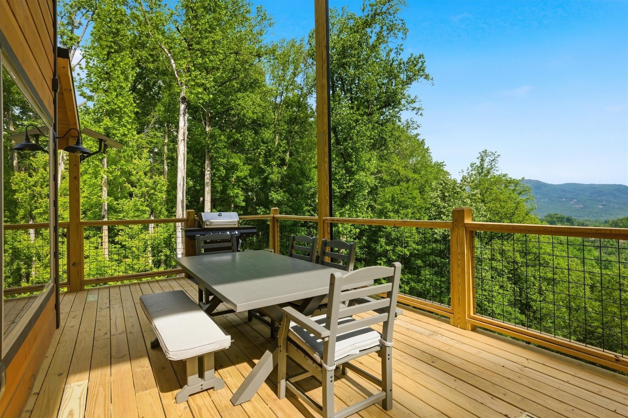 Um deck de madeira com mesa, cadeiras, churrasqueira e vista para uma área arborizada.