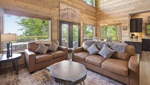 A spacious living room with a high ceiling, wooden walls, and large windows offering a view of greenery. - Our Together Place 3 Bedroom Cabin (Sevierville)