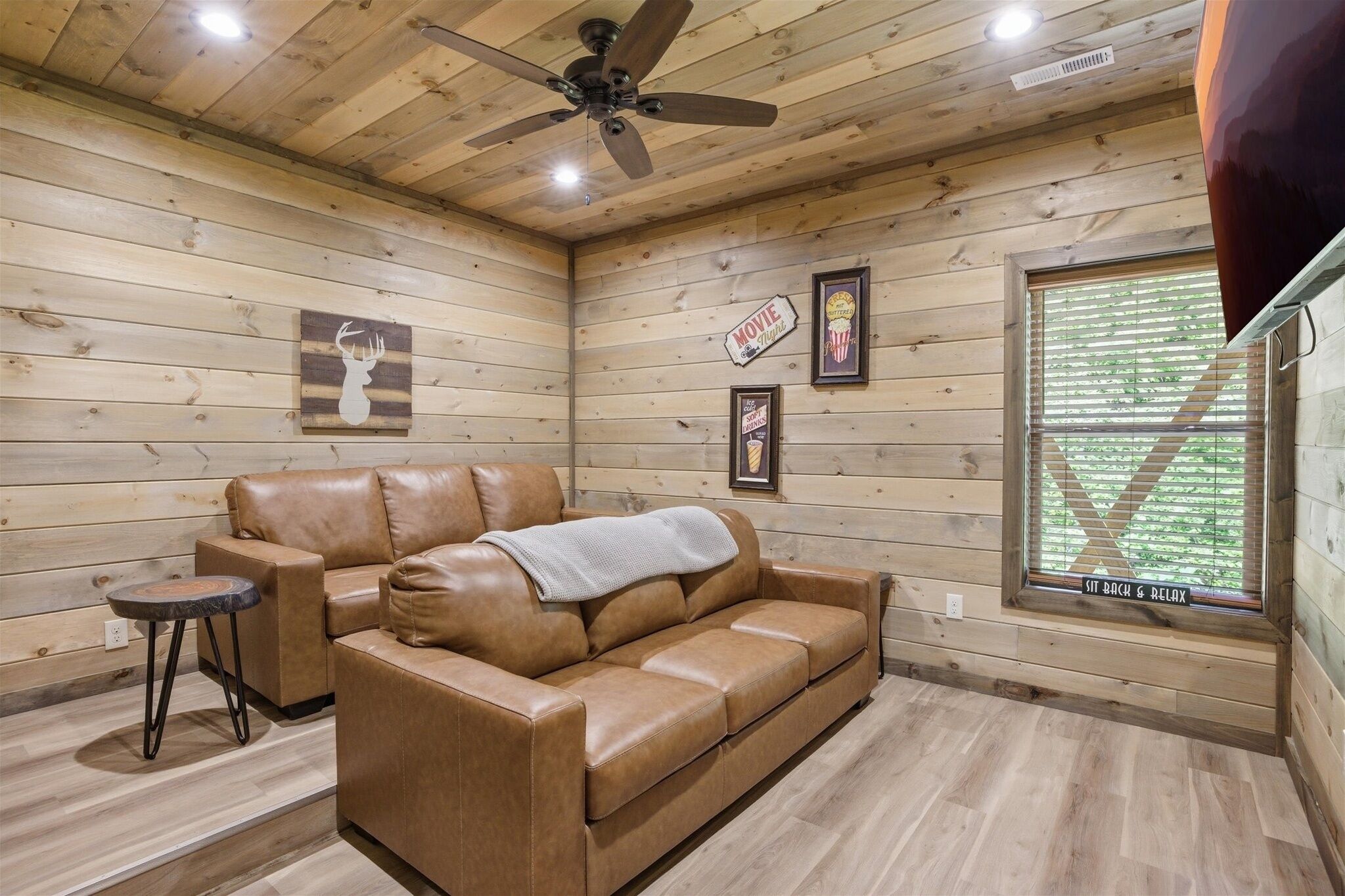 A wooden interior with a leather sofa, a ceiling fan, framed movie posters, and a window with blinds.