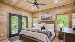 A wooden cabin bedroom with a bed, a ceiling fan, a nightstand, and a window with a view of greenery. - Our Together Place 3 Bedroom Cabin (Sevierville)