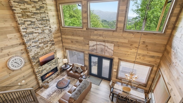 A wooden interior with a stone fireplace, a sofa, a dining table, and large windows offering a view of greenery. - Our Together Place 3 Bedroom Cabin (Sevierville)