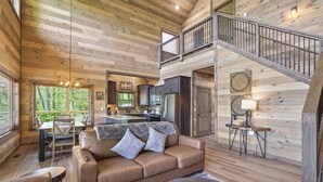 A spacious living area with a high ceiling, wooden walls, a sofa, a dining table, and a staircase. - Our Together Place 3 Bedroom Cabin (Sevierville)