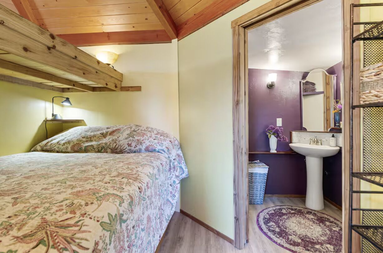 A bedroom with a bed, a bedside lamp, a wooden ceiling, and a view into a bathroom with a sink and mirror.