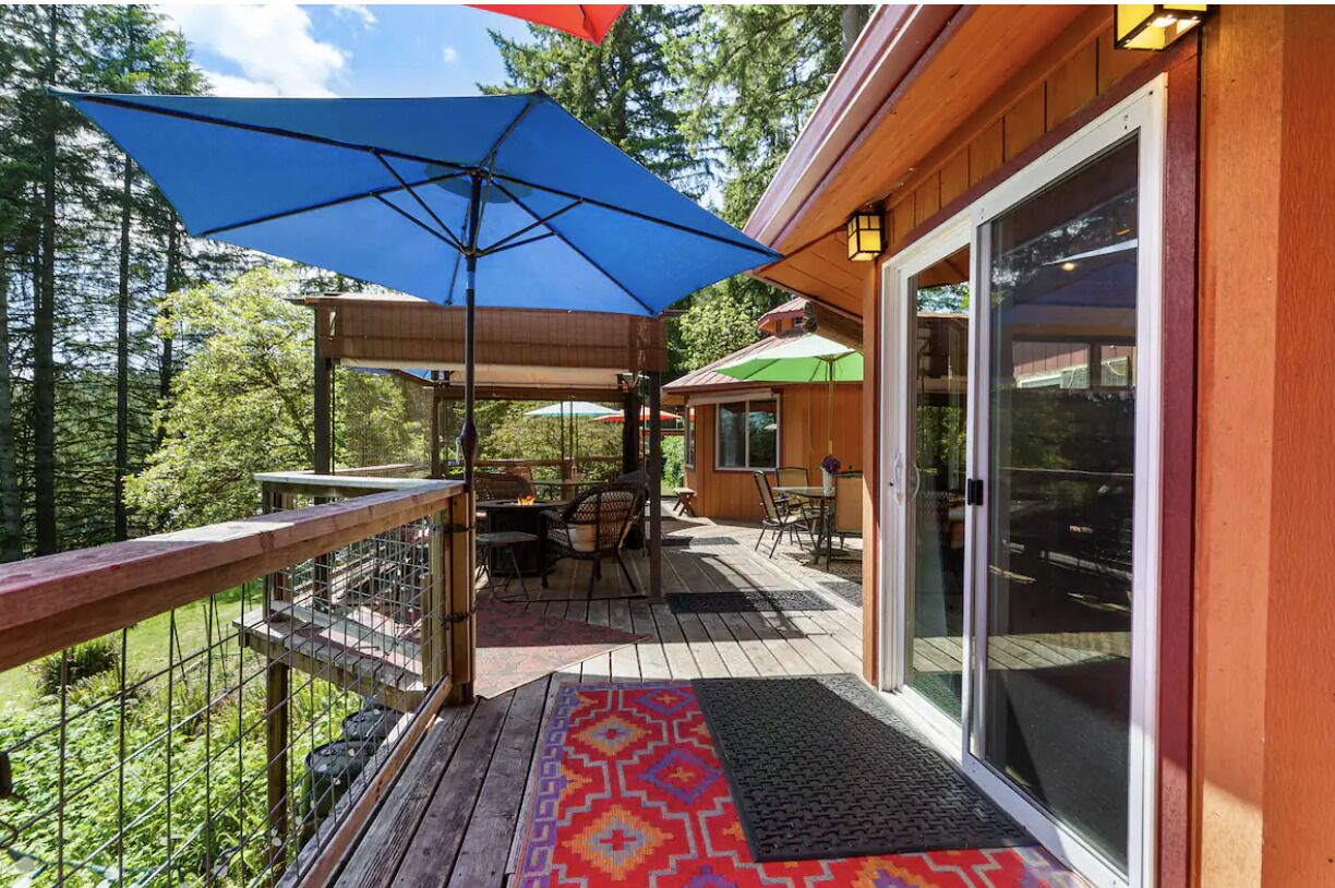 Une terrasse en bois avec des meubles d’extérieur, un parasol bleu et une porte vitrée donnant sur un espace abrité.
