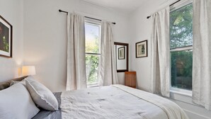 Une chambre à coucher avec un lit, deux fenêtres avec des rideaux, une table de chevet avec une lampe et des tableaux encadrés accrochés au mur.